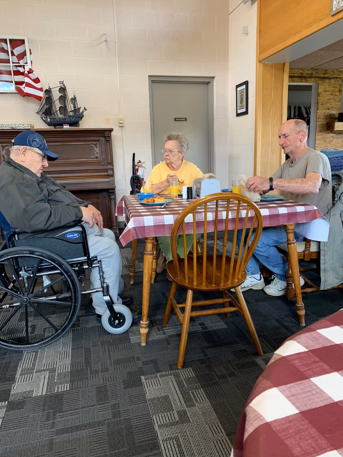 Three older adults sitting around a red-and-white checkered dining table in a communal dining room, with a wheelchair, piano, and an American flag in the background.