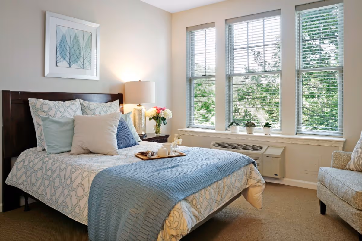 A bright furnished bedroom with a made bed, bedside lamp and tray, large windows with blinds and potted plants.