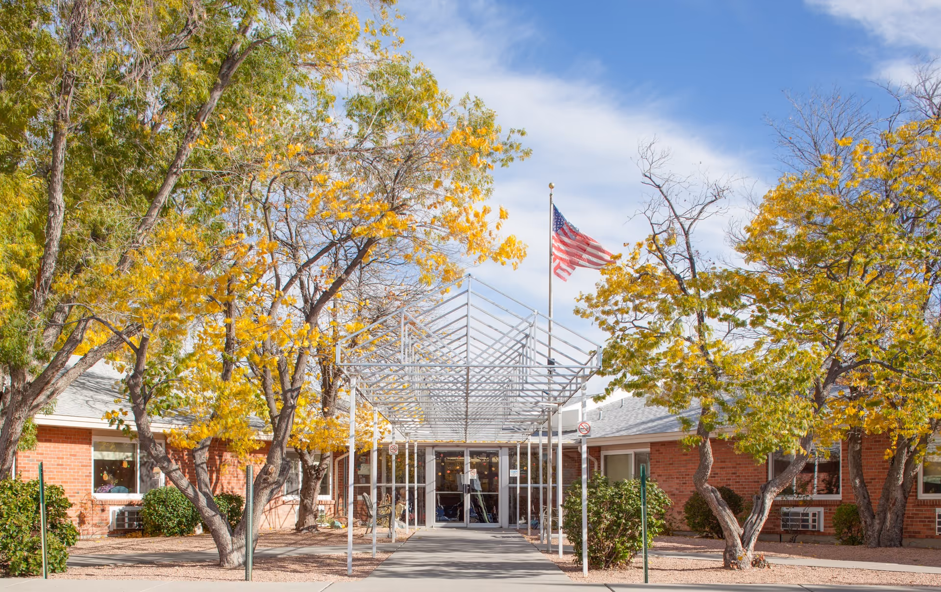 Entrance of a brick building with a covered walkway supported by white metal poles. Trees with yellow leaves surround the entrance, and an American flag is flying on a flagpole above the walkway. The sky is partly cloudy.