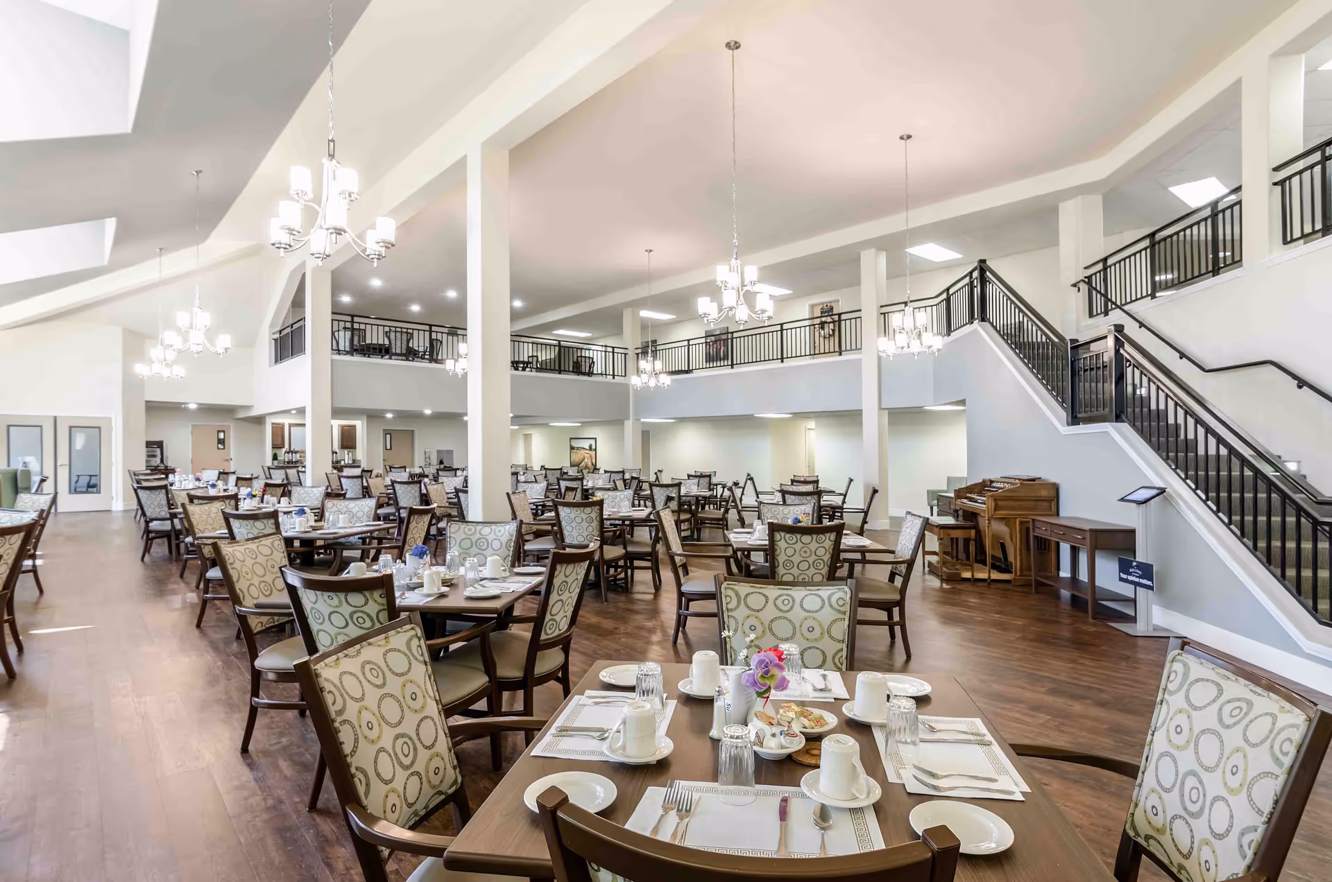 Spacious dining room in a senior living facility with multiple tables and chairs arranged neatly. Each table is set with white cups, plates, utensils, and napkins. The room features high ceilings with hanging chandeliers, a wooden floor, and a staircase leading to an upper level with railings. There is a piano and a small table near the staircase.
