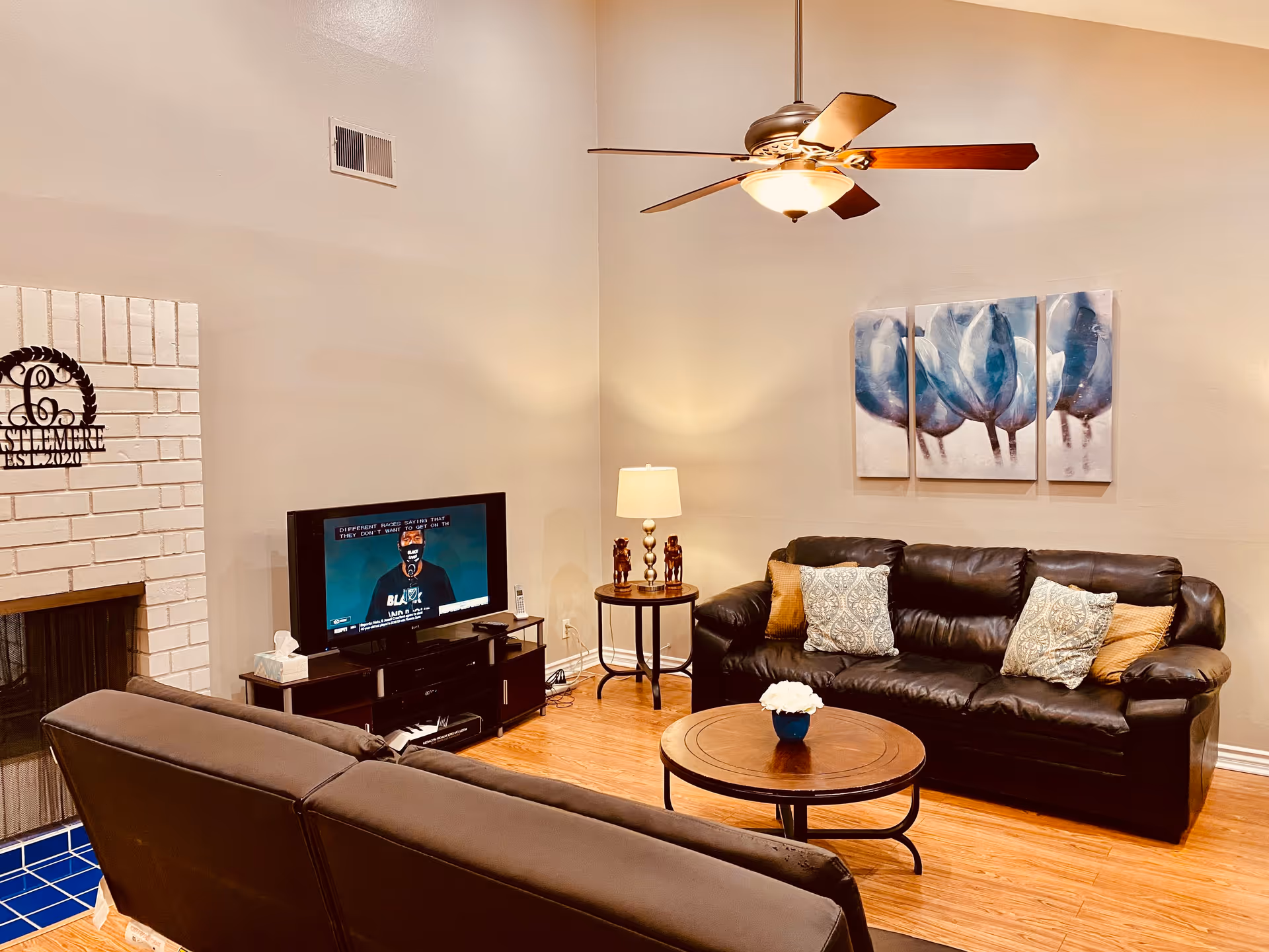 A cozy living room with two dark leather sofas, a round wooden coffee table with a small flower pot, a TV on a stand, a side table with a lamp and decorative items, a ceiling fan with light, and a white brick fireplace with a Castlemere sign above it. The walls are beige and there is a three-panel blue floral painting hanging above one sofa.