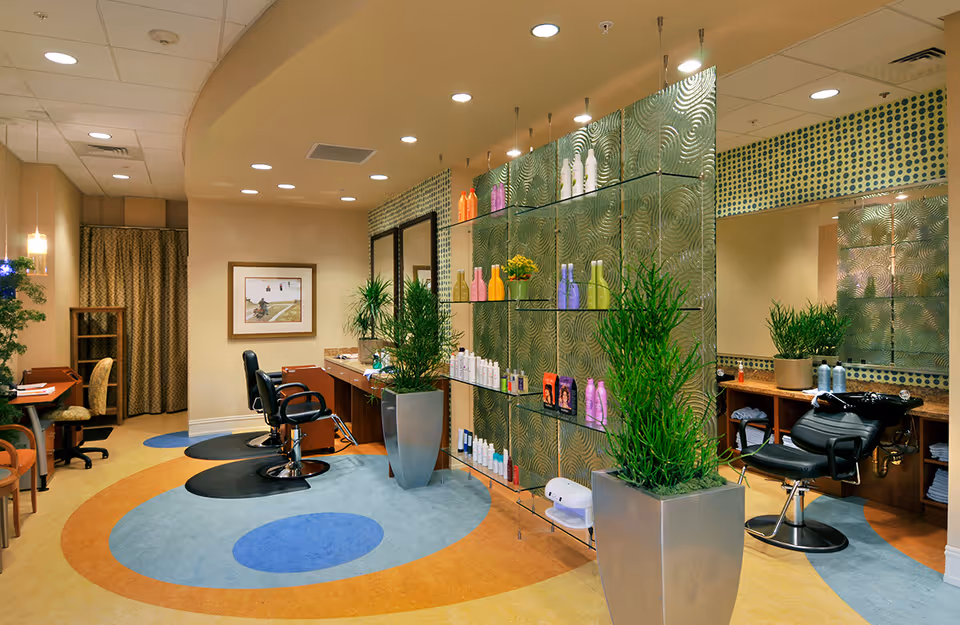 Interior view of a salon area in a retirement community featuring salon chairs, mirrors, shelves with hair care products, potted plants, and a colorful circular patterned floor.