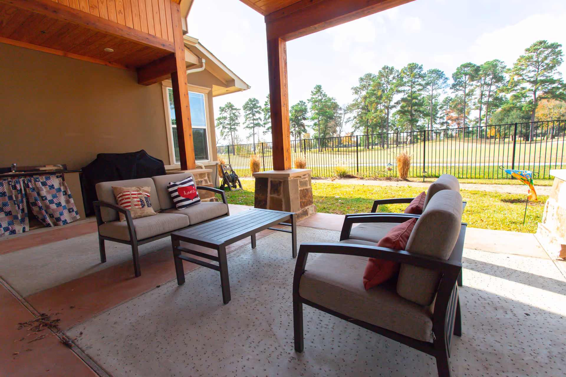 Covered outdoor patio area with cushioned seating including two loveseats and a coffee table. The patio has wooden beams and overlooks a fenced grassy area with trees in the background. There is a grill covered with a black cover and a small counter with a patriotic-themed cloth on the left side.