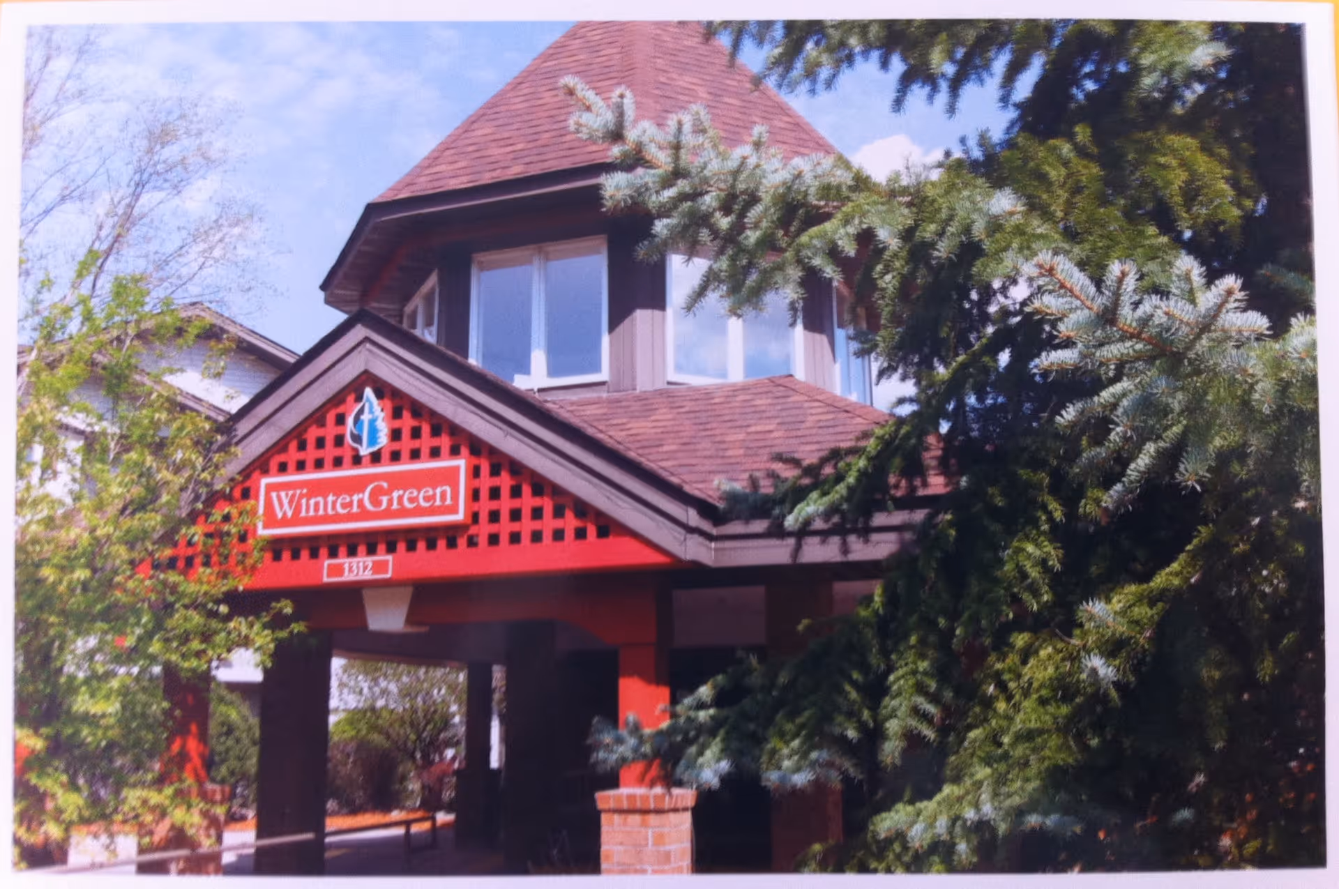 Exterior view of WinterGreen Independent Senior Living facility entrance with a red roofed structure and surrounding trees under a blue sky.