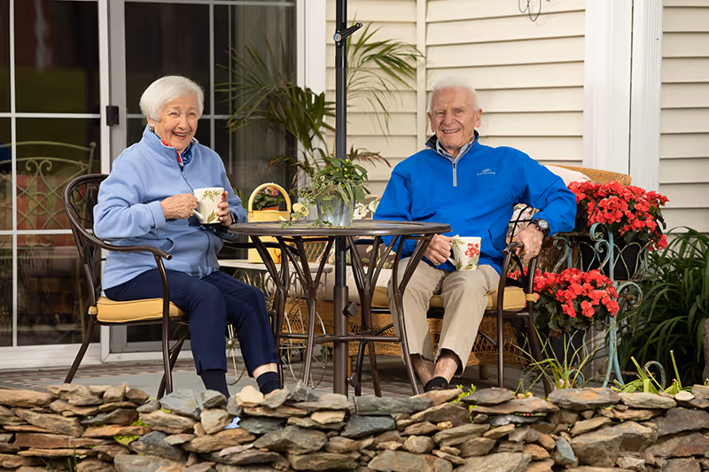 An elderly woman and man sitting outside on a patio at a round metal table with a glass top, each holding a floral-patterned mug. They are smiling and appear to be enjoying a pleasant moment. The patio is decorated with potted red flowers and greenery, and there is a stone wall in the foreground. The background shows a sliding glass door and the exterior siding of a building.