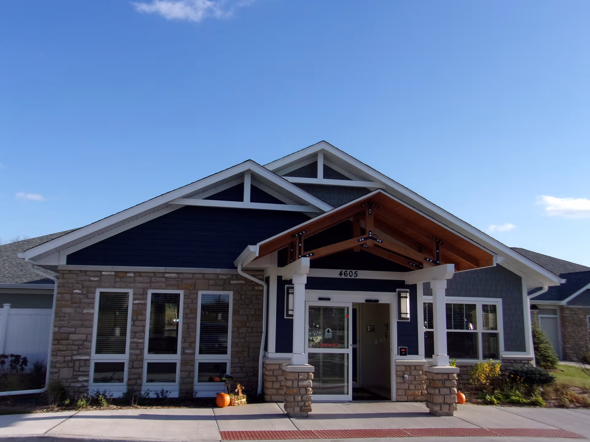 Front exterior view of a single-story building with stone and blue siding, a covered entrance with wooden beams, and the number 4605 above the door. There are pumpkins and plants near the entrance, under a clear blue sky.