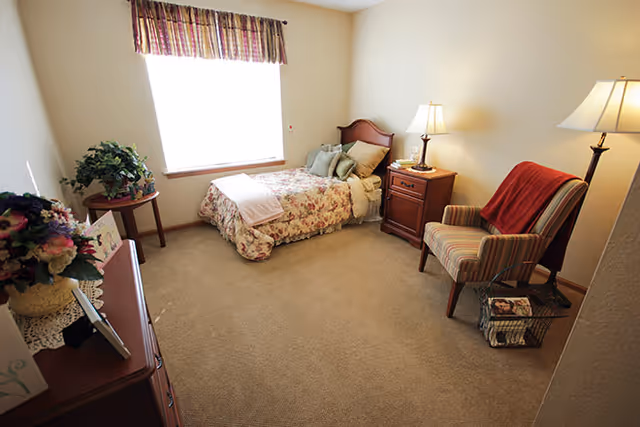 A cozy bedroom with a single bed covered in floral bedding, a wooden nightstand with a lamp, a striped armchair with a red throw blanket, a small table with a plant, and a dresser with flowers and framed pictures. The room has beige walls and carpet, and a window with a valance letting in natural light.