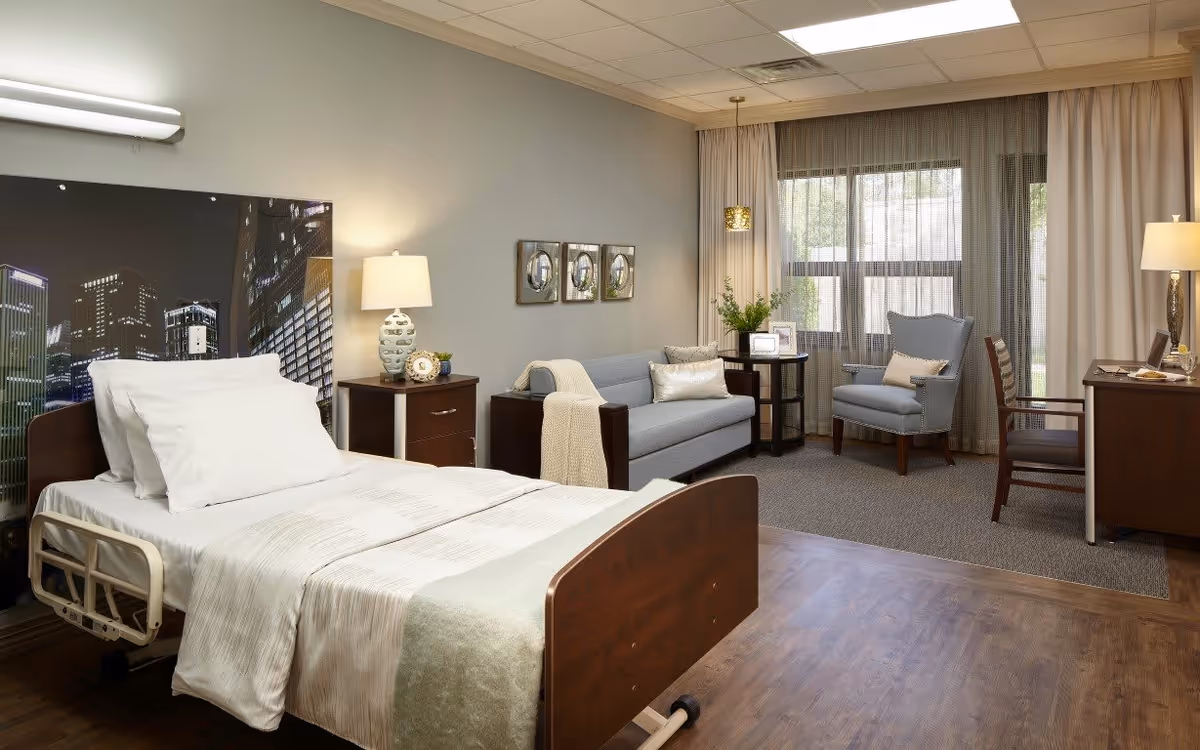 A well-lit senior living bedroom featuring a hospital-style bed with white linens and a cityscape headboard. The room includes a wooden nightstand with a lamp and clock, a gray sofa with a throw blanket and pillows, a small round table with framed photos and a plant, two armchairs, a wooden desk with a lamp, and large windows with sheer and opaque curtains allowing natural light.