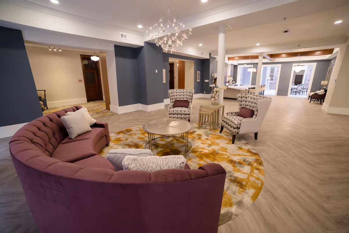 Spacious modern common lounge with a curved purple sofa, patterned armchairs, round coffee table, and chandelier.