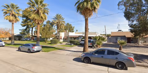 Street view of a residential area with palm trees and parked cars in front of single-story buildings under a clear blue sky.