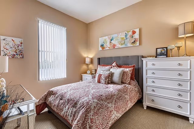 Well-lit bedroom with a double bed dressed in patterned bedding, a white dresser, nightstands, lamps, and wall art.