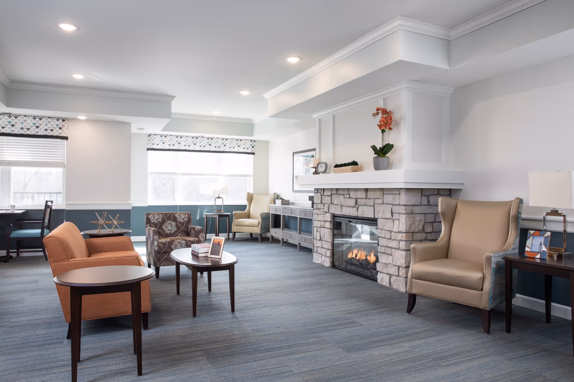 A bright and spacious living room area with a stone fireplace featuring a lit fire. The room has a mix of comfortable seating including an orange armchair, a floral patterned armchair, and a beige wingback chair. There are several wooden side tables and a console table against the wall. Large windows with patterned valances allow natural light to fill the room.