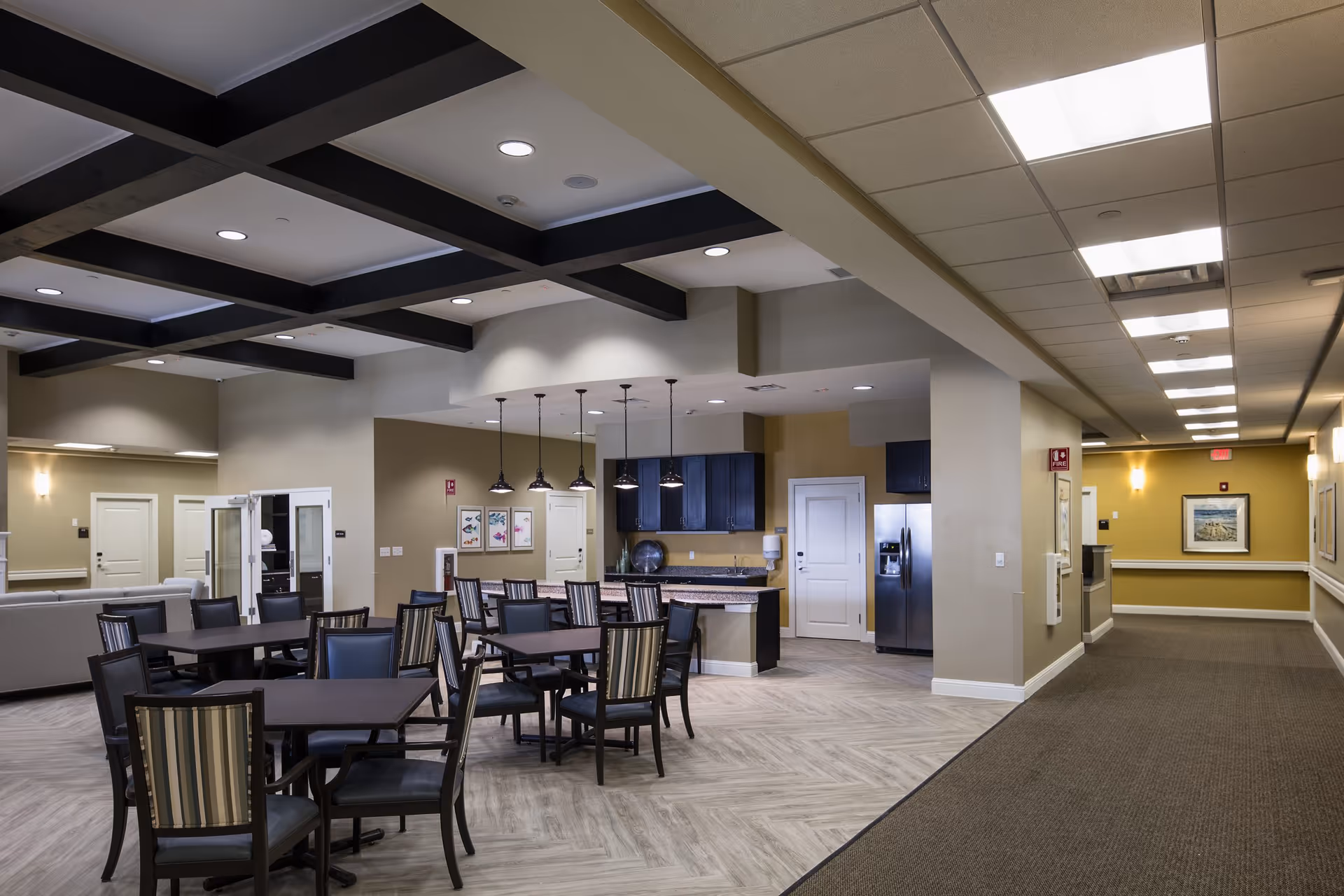 Interior view of a memory care facility dining and common area with multiple tables and chairs, a kitchen area with hanging pendant lights, dark cabinets, and a stainless steel refrigerator. The space has a mix of carpet and wood-patterned flooring, beige walls, and a ceiling with recessed lighting and exposed beams.