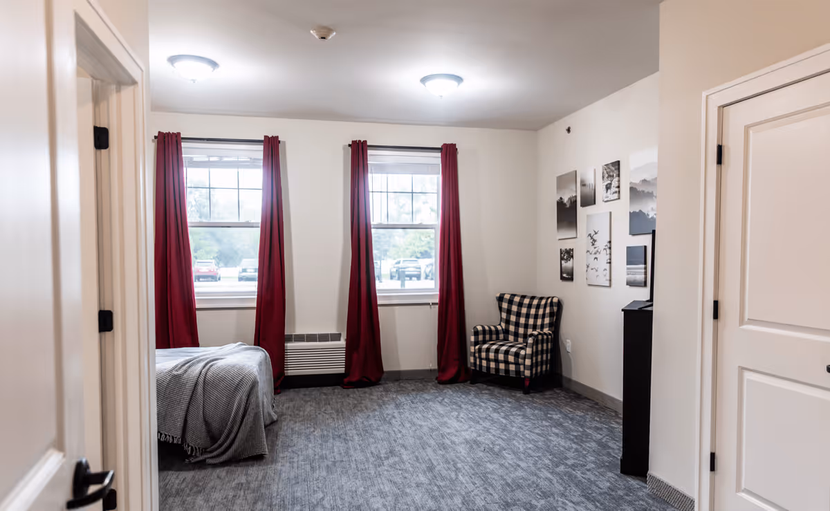 A senior living facility bedroom with two large windows covered by red curtains, a bed partially visible on the left side with a gray blanket, a black and white checkered armchair in the corner, and several framed pictures on the wall. The room has gray carpet flooring and white walls with two ceiling lights.