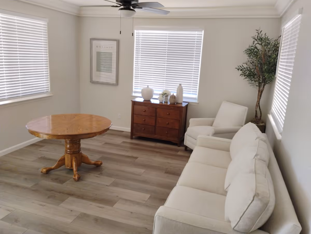 Bright, minimally furnished living room with a beige sofa and armchair, a round wooden table, a small wooden dresser beneath windows, and a potted plant.