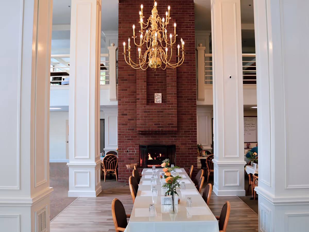 A dining room with a long table set with placemats, glasses, and floral centerpieces. The room features a large brick fireplace with a fire burning and a gold chandelier hanging from the ceiling. White columns and a second-floor balcony are visible in the background.