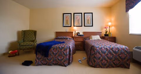 A warmly lit bedroom with two twin beds separated by a nightstand with lamps, a green armchair, and a window.