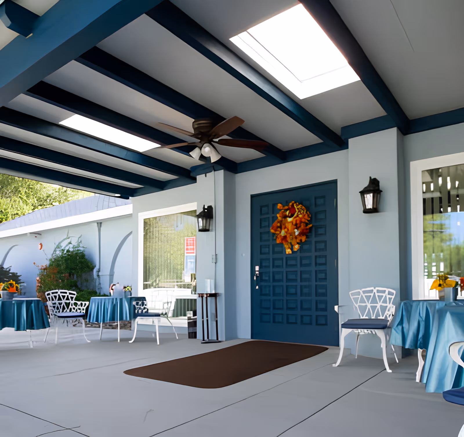 Covered outdoor patio area with a blue ceiling featuring exposed beams and skylights, a ceiling fan, a blue door decorated with an orange wreath, white chairs with blue cushions, and tables covered with blue tablecloths. There are wall-mounted lantern-style lights and some greenery visible outside.