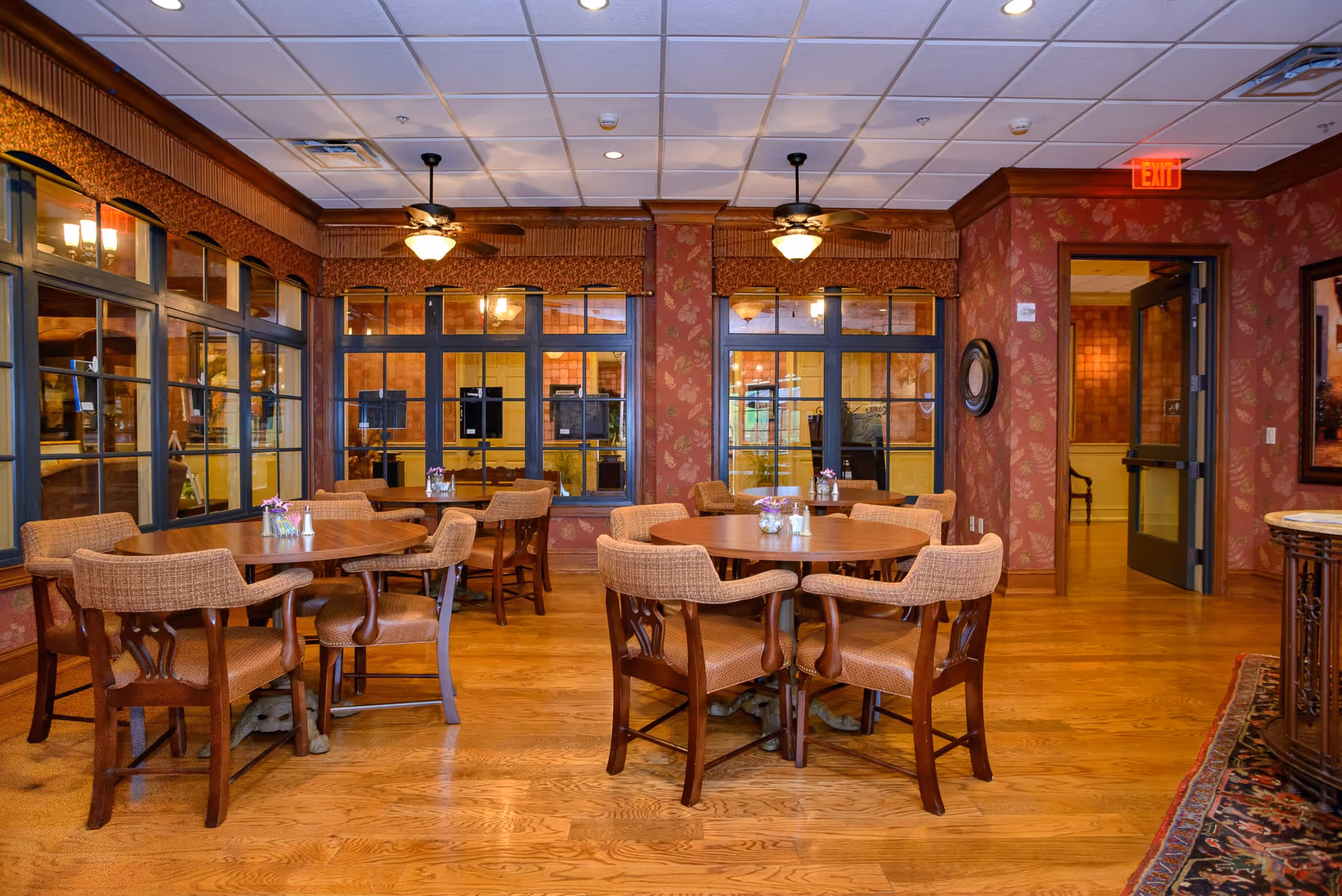 Interior view of a dining room with round wooden tables and cushioned chairs. The room has wooden flooring, patterned wallpaper, ceiling fans with lights, and large windows with dark frames. There is an exit door on the right side and decorative elements including a framed picture and a round mirror on the walls.