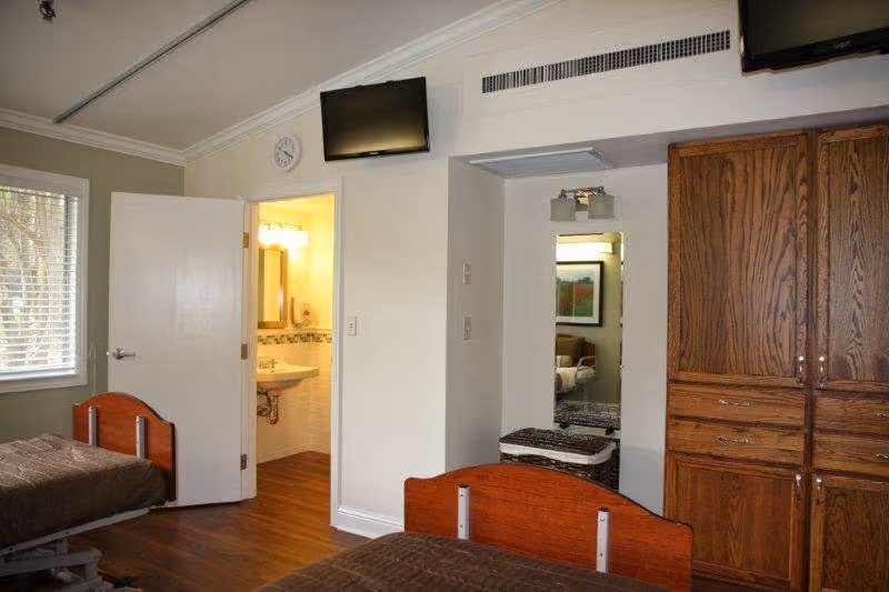 Two-bed resident bedroom with wooden headboards, an open bathroom door, a wall-mounted TV, and a wooden wardrobe.