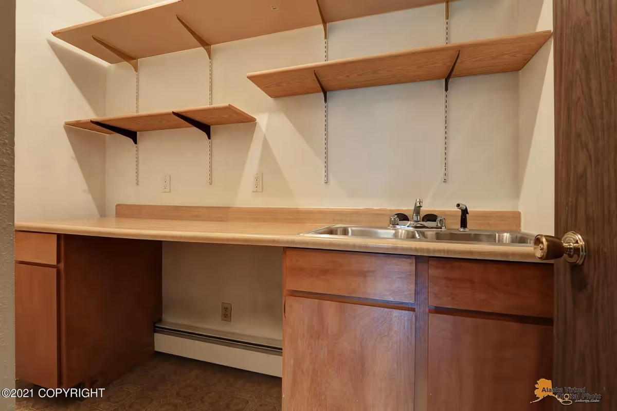 A small room with a countertop featuring a double stainless steel sink and wooden cabinets below. Above the countertop, there are multiple wooden shelves mounted on the wall. The room has a beige wall and a brown door with a brass doorknob partially visible on the right side.