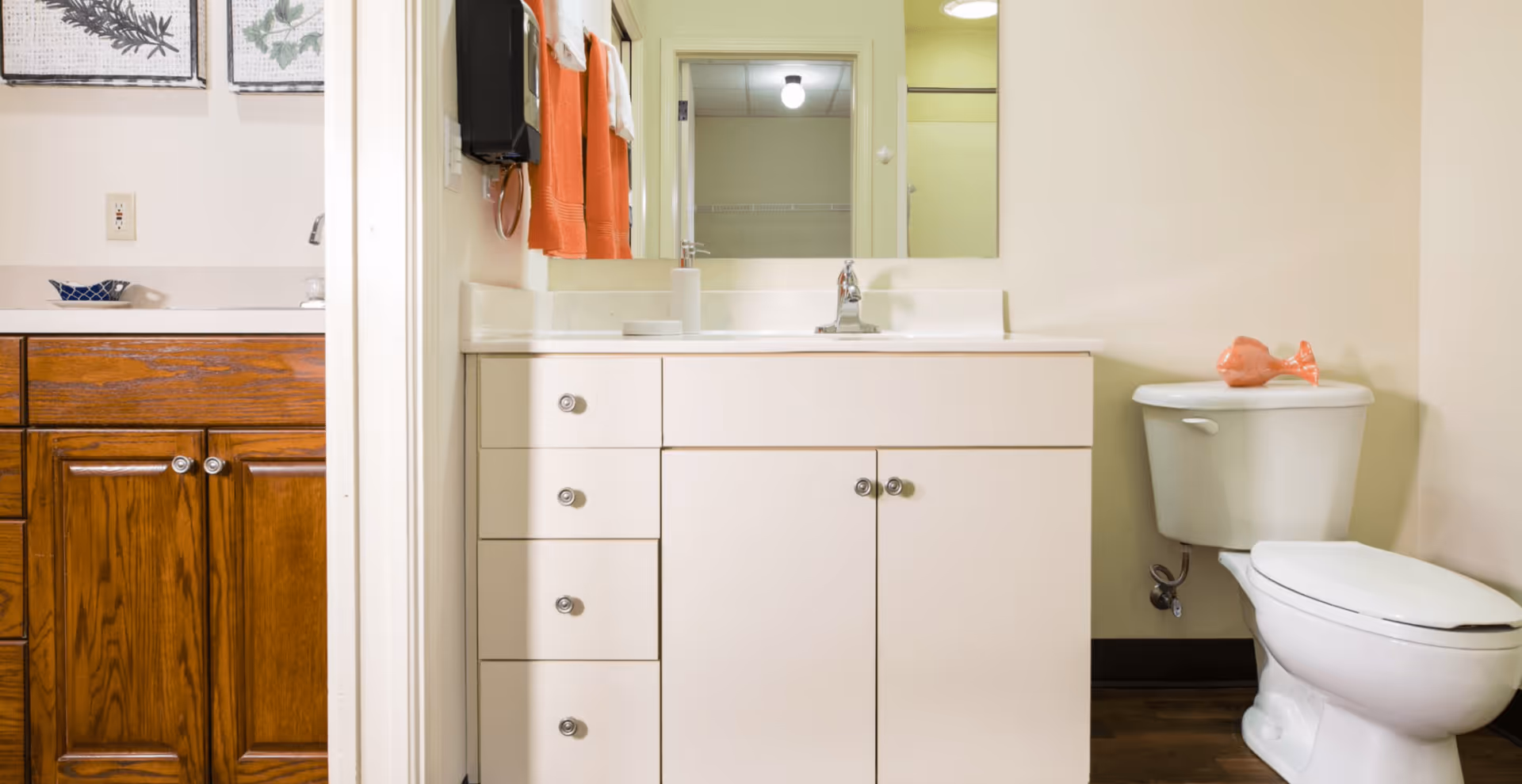 A bathroom with a white vanity cabinet and sink, a mirror above the sink, orange towels hanging on the wall, and a white toilet with a small orange decorative fish on the tank lid. Adjacent to the bathroom is a glimpse of a kitchen area with wooden cabinets and a countertop.