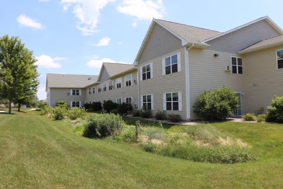 Exterior view of a two-story beige senior living facility building with multiple windows, surrounded by green grass, bushes, and a tree under a partly cloudy blue sky.
