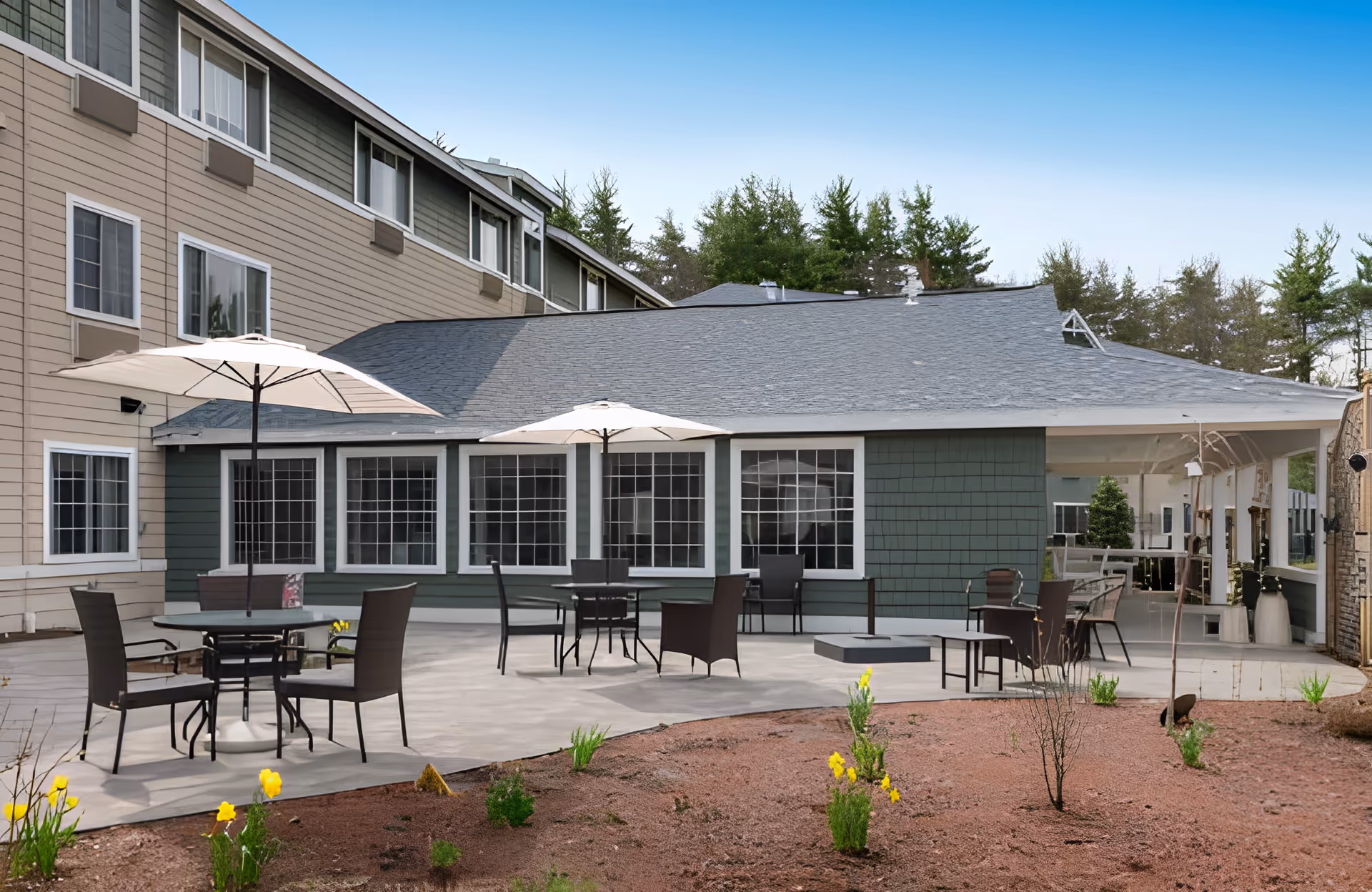 Outdoor patio area at Ledgewood Bay Assisted Living with several tables and chairs under large white umbrellas, adjacent to a building with multiple windows and surrounded by a garden with small plants and yellow flowers.