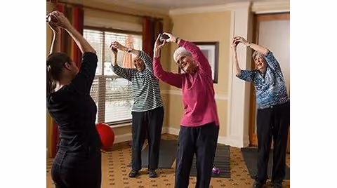 A group of three elderly women and an instructor participating in a stretching exercise inside a room with carpeted floor and large windows with curtains.