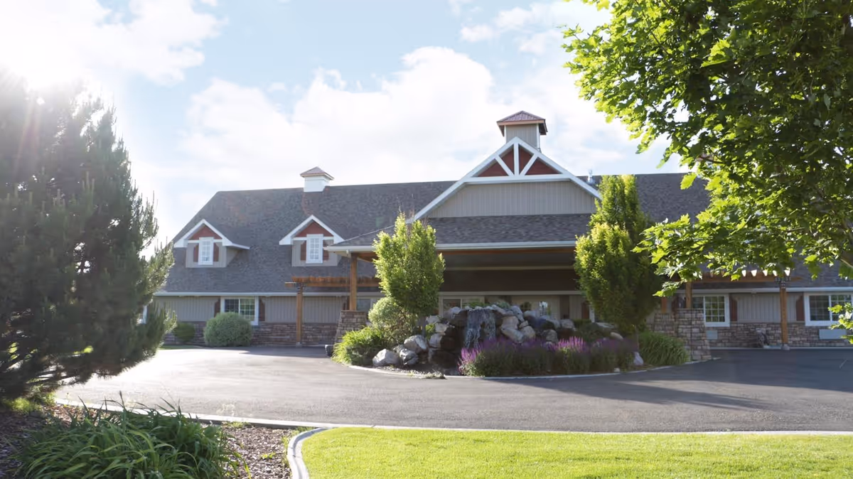 Exterior view of a large assisted living facility building with a peaked roof, dormer windows, and a covered entrance. The building is surrounded by well-maintained landscaping including trees, bushes, and a rock garden with purple flowers. The sky is partly cloudy with sunlight shining through.