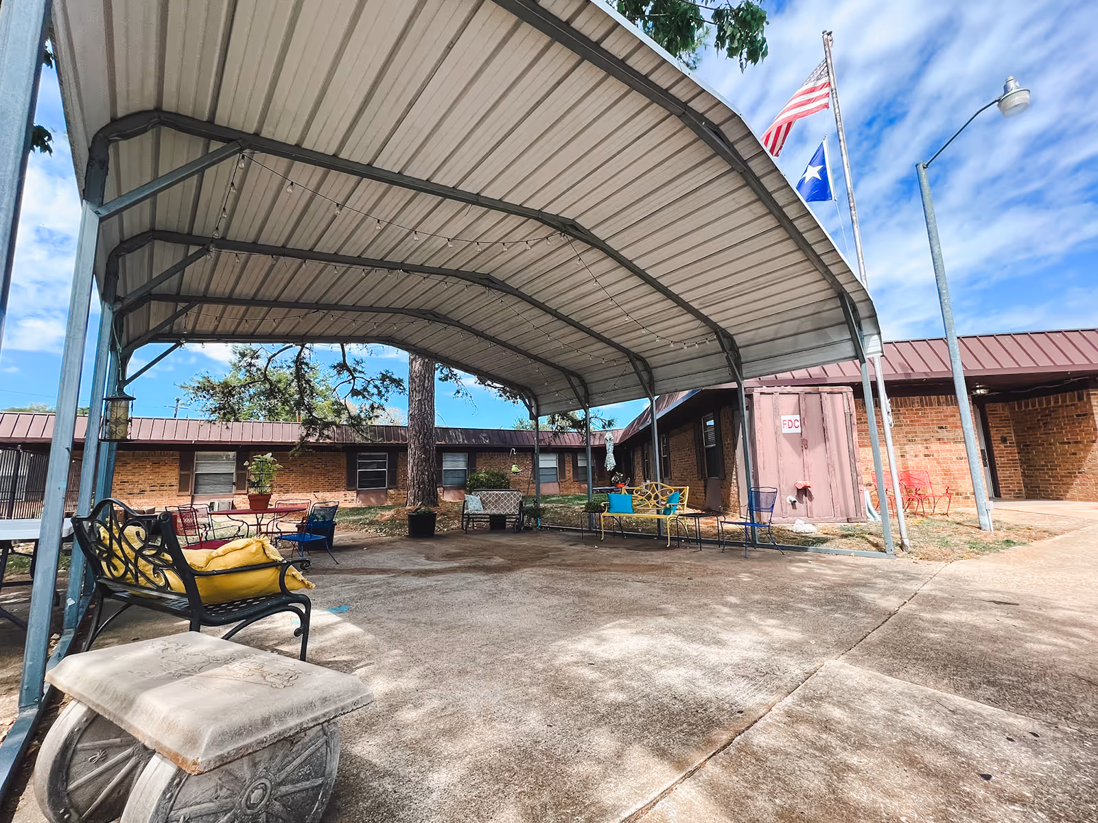 Outdoor covered seating area with metal benches and chairs, some with yellow and blue cushions, under a large metal canopy. The area is surrounded by a brick building with windows and a door, and two flagpoles flying the American and Texas flags. The sky is partly cloudy.