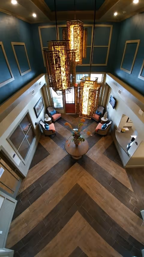 Overhead view of a two-story lobby with large hanging pendant lights, a central round table with flowers, seating alcoves and a chevron-pattern wood floor.