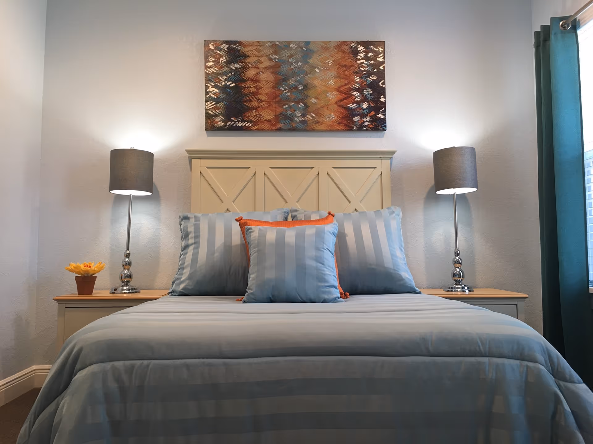 Neatly made bed with striped blue bedding and decorative pillows, a headboard with a colorful painting above, and two bedside lamps.
