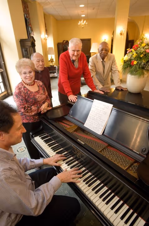 A group of four elderly people standing around a grand piano while a man plays it. The setting appears to be a warmly lit common area with yellow walls, decorative lighting, and a large floral arrangement on the piano.