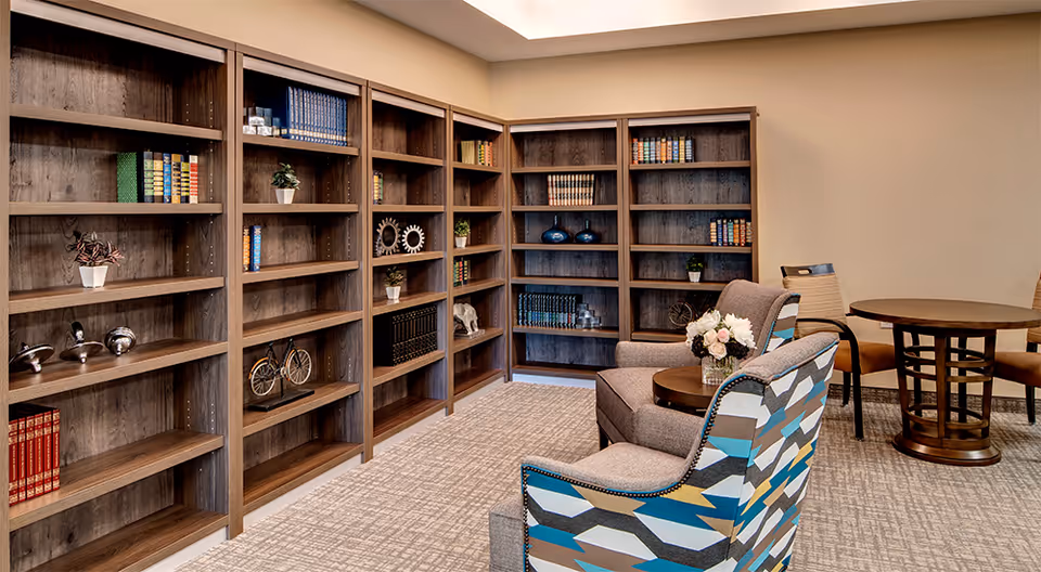 A cozy reading or sitting area in a senior living community featuring wooden bookshelves filled with books and decorative items, two upholstered armchairs with a small round table between them holding a vase of flowers, and a round wooden table with chairs in the background.