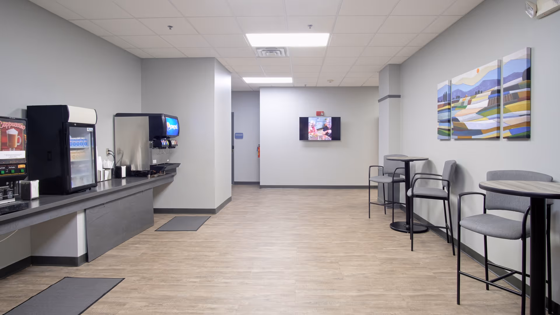 A clean and modern common area with a beverage station on the left side featuring a coffee machine, a soda dispenser, and a refrigerator. On the right side, there are high tables with chairs and a three-panel abstract landscape painting on the wall. The floor is light wood, and the walls are painted light gray. A small TV is mounted on the far wall.