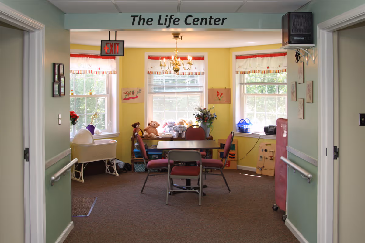 A bright activity room labeled "The Life Center" with a table and chairs, windows, and stuffed animals on a shelf.