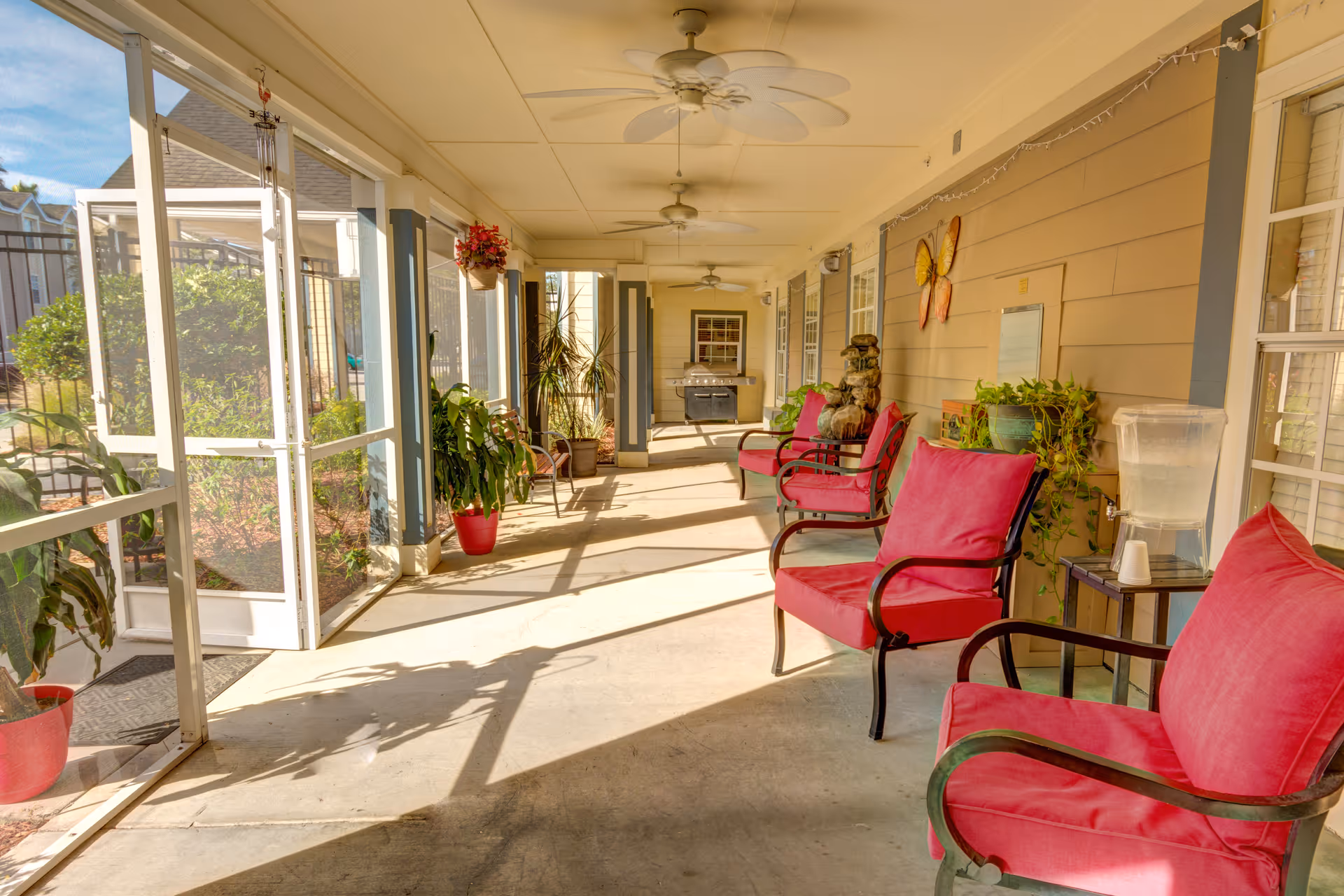 A bright and spacious screened-in porch area with several red cushioned chairs arranged along the wall. The porch has ceiling fans, potted plants, a water dispenser on a small table, and a barbecue grill at the far end. The area is well-lit with natural sunlight and overlooks a garden outside.