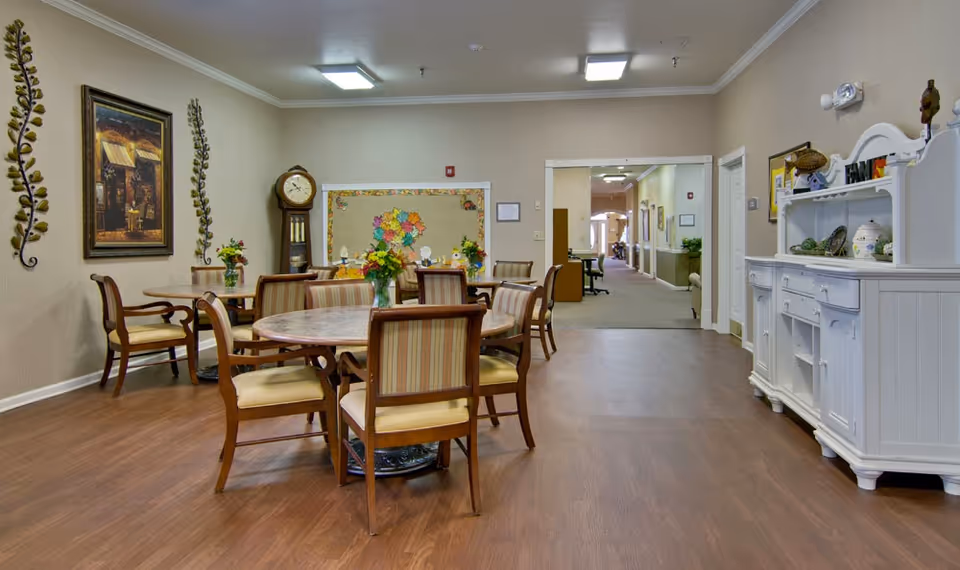 A well-lit common area with round tables and wooden chairs with striped cushions. The room has wooden flooring, beige walls, and decorative wall art including a large clock and floral arrangements on the tables. A white cabinet with decorative items is on the right side, and a hallway is visible in the background.