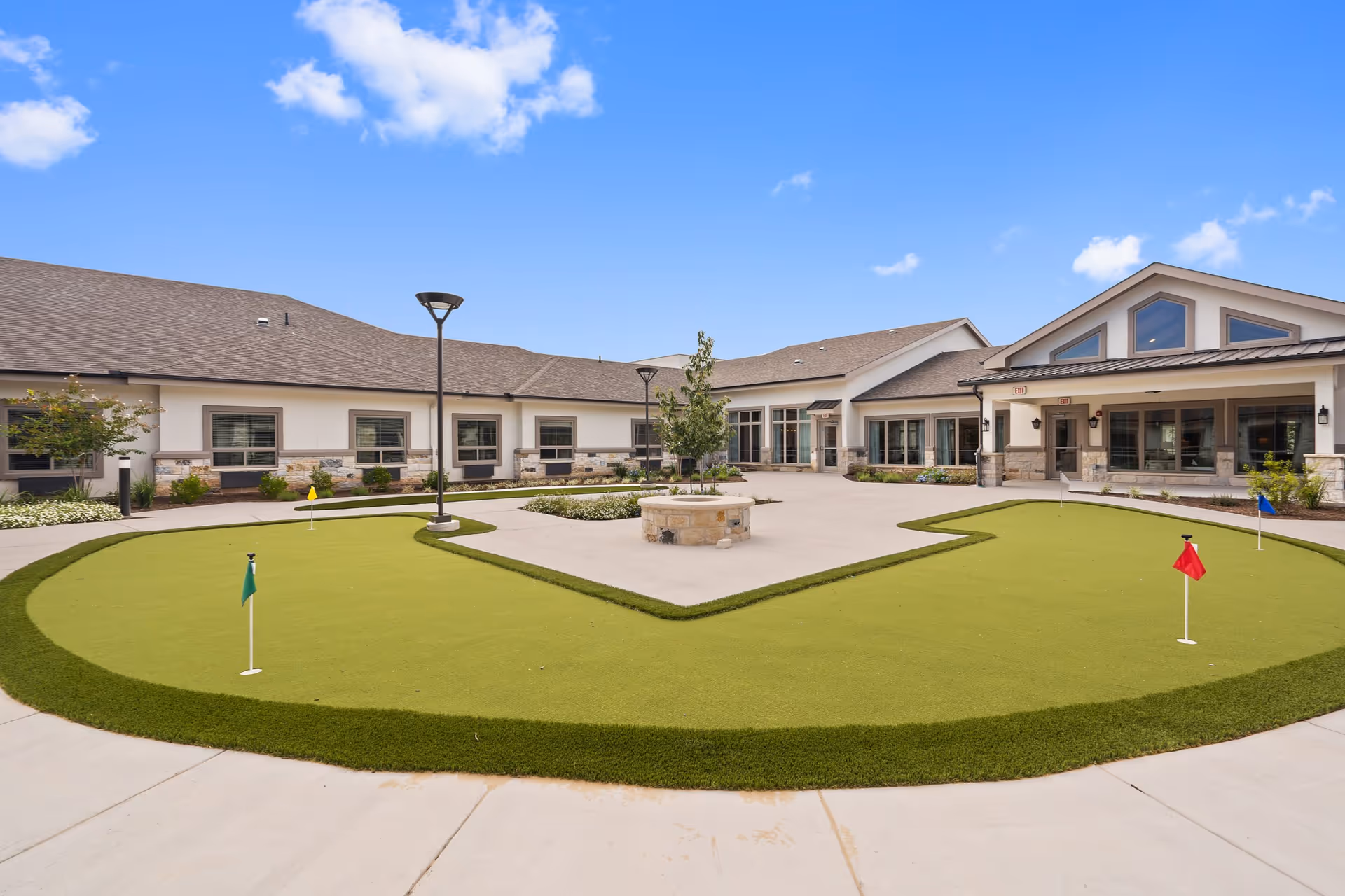 Outdoor courtyard area at Volante Senior Living of Sage Spring featuring a putting green with multiple colored flags, a central stone fire pit, surrounding landscaping, and a building with large windows under a clear blue sky.