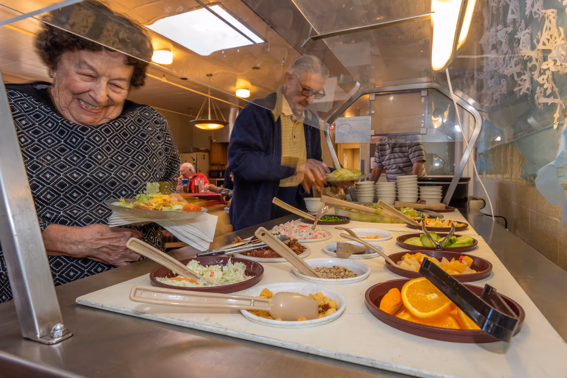 Two elderly individuals serving themselves food from a salad bar in a dining area. Various salad ingredients and fruit slices are visible in bowls on the counter, with a protective glass barrier above the food. The setting appears warm and welcoming with other people seated in the background.