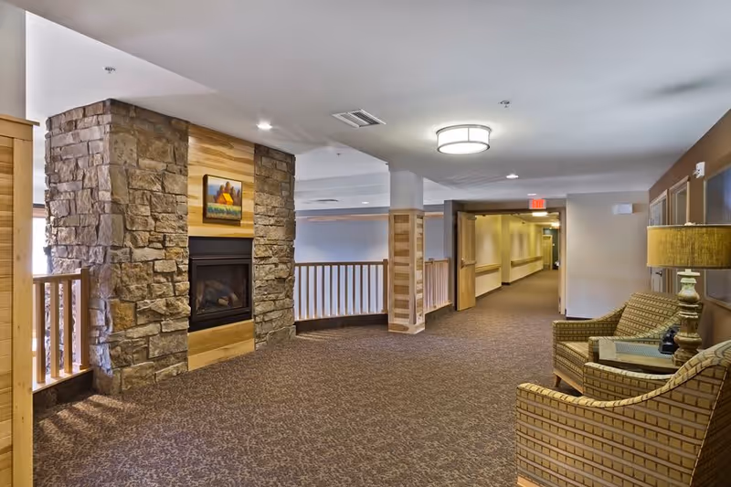 Interior hallway of a senior living facility featuring a stone and wood fireplace on the left, two patterned armchairs with a side table and lamp on the right, and a carpeted floor leading to an open doorway and corridor with handrails.