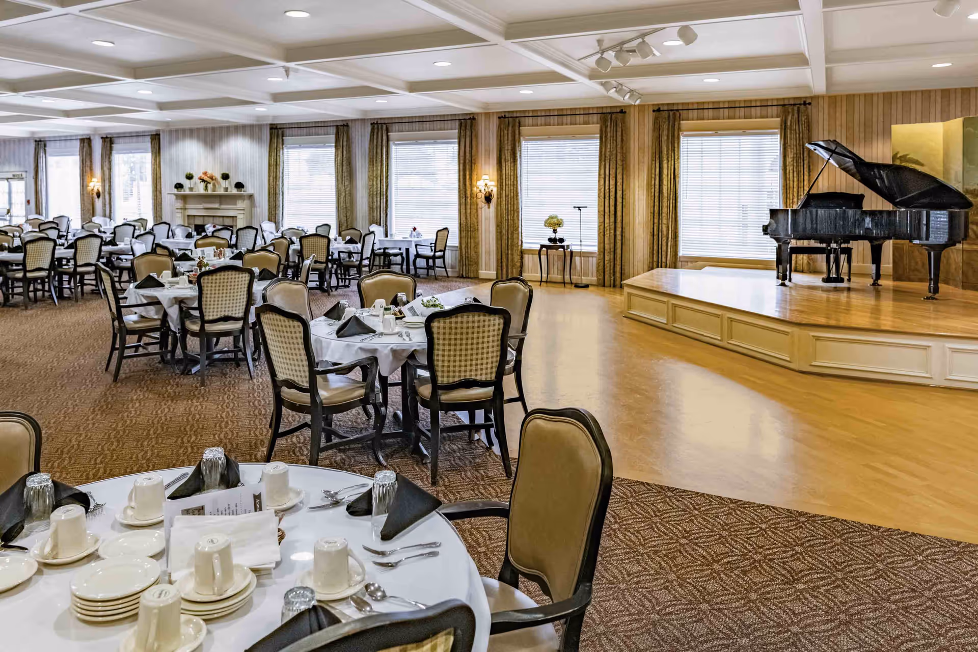 Spacious dining room with round tables set for a meal and a black grand piano on a raised stage.