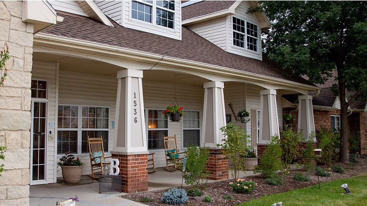 Front porch of a residential building with beige siding and brick pillars, featuring rocking chairs, potted plants, hanging flower baskets, and the number 1536 displayed on one pillar. The porch is surrounded by a landscaped garden with shrubs and flowers.