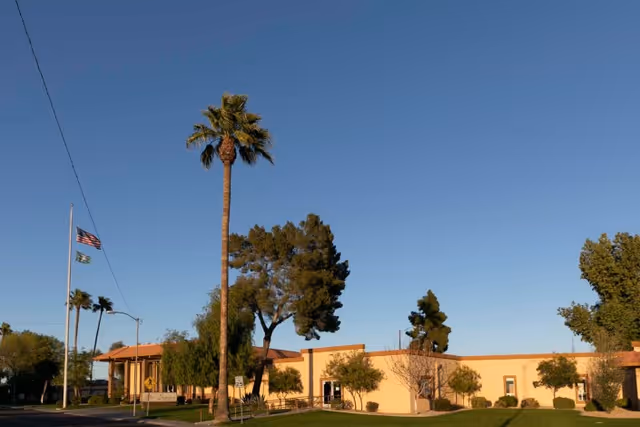 Exterior view of Sunview Health And Rehabilitation Center building with a clear blue sky, several trees including a tall palm tree, a flagpole with the American flag and another flag, and a well-maintained lawn in front.