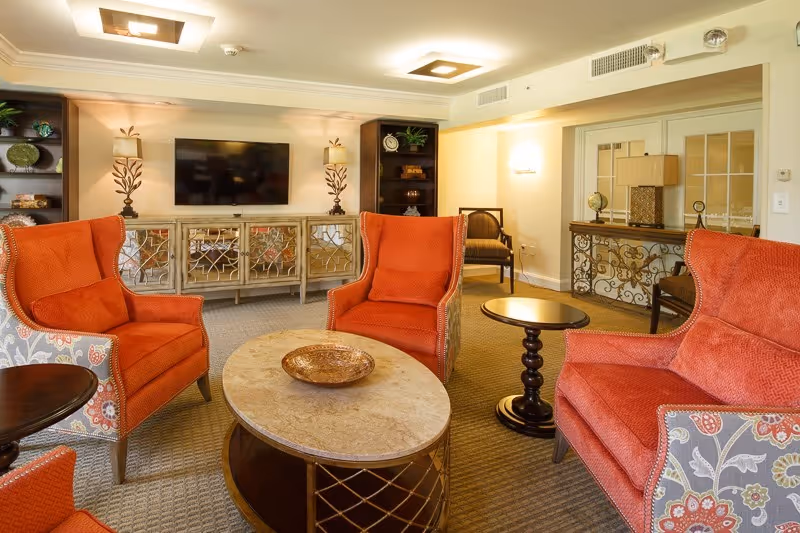 A cozy living room area with four orange upholstered armchairs arranged around a marble-top coffee table with a decorative bowl. Behind the chairs is a mirrored cabinet with a flat-screen TV mounted on the wall above it, flanked by two decorative lamps. The room has warm lighting, carpeted floors, and additional seating and furniture along the walls.