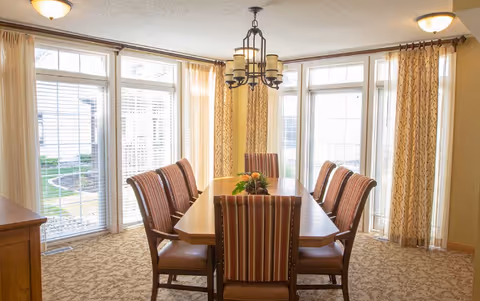 A dining room with a rectangular wooden table surrounded by eight striped upholstered chairs. The room has large windows with white blinds and beige curtains, allowing natural light to fill the space. A decorative chandelier hangs above the table, and there is a small centerpiece with flowers on the table. The floor is carpeted with a patterned design.