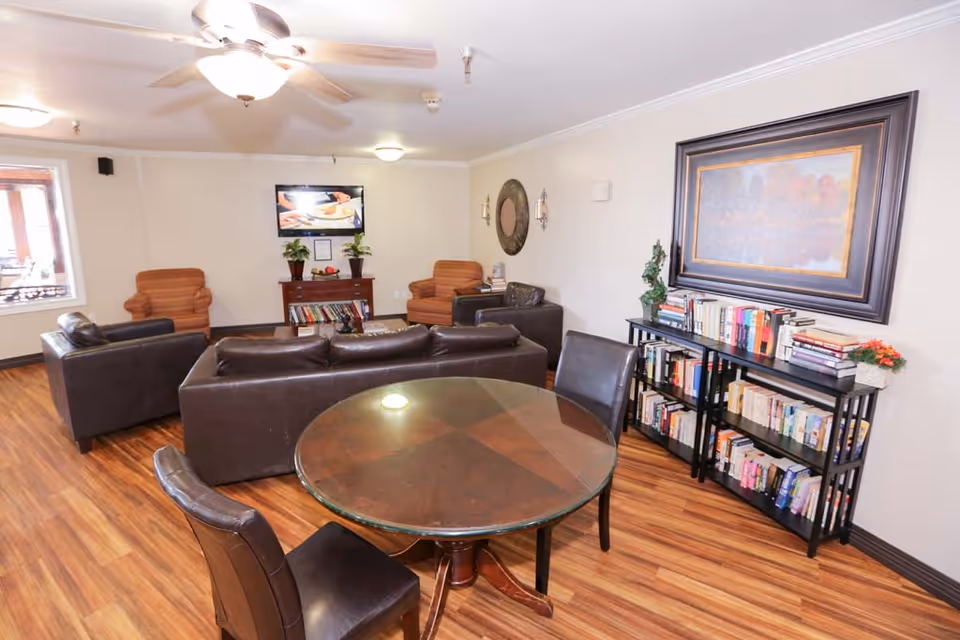 A cozy living room area with wooden flooring, featuring a round glass-top table with four dark chairs in the foreground. Behind the table are two dark leather sofas and two orange armchairs arranged around a small wooden cabinet with plants and books. A bookshelf filled with books and a large framed painting are on the right wall. The room is well-lit with ceiling lights and a ceiling fan.