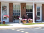 Outdoor patio area of Westbrook Care Center with two chairs, a bench, flower pots with pink flowers, and a small statue of a child. The building has a brick exterior with two windows and two doors.