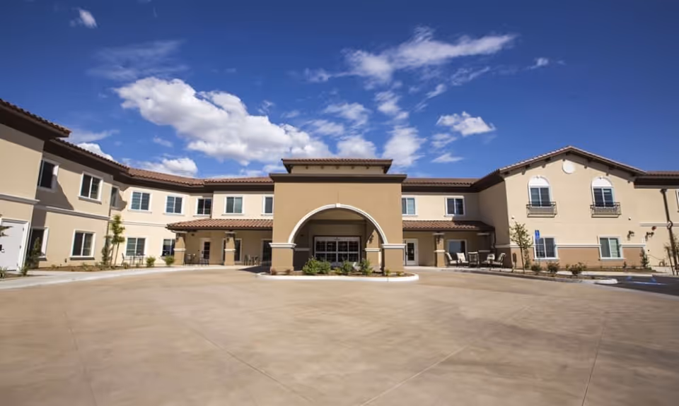 Front exterior view of a two-story senior living facility building with beige walls, brown roof tiles, and an arched entrance under a blue sky with scattered clouds.